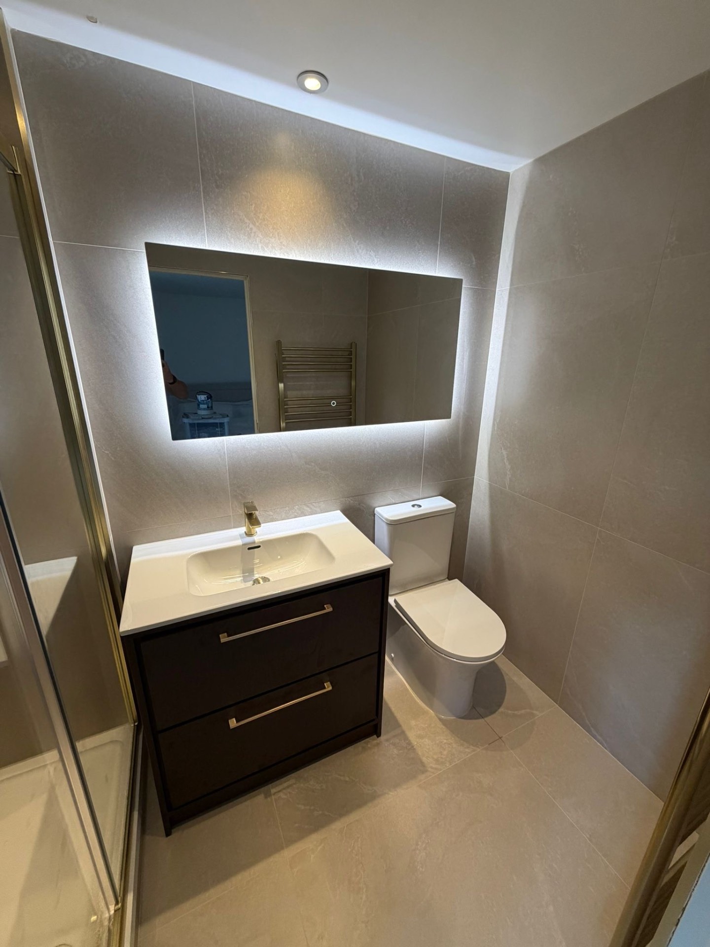 Grey-tiled cloakroom with dark wood vanity, LED backlit mirror and brushed brass fittings — bathroom renovation Basingstoke